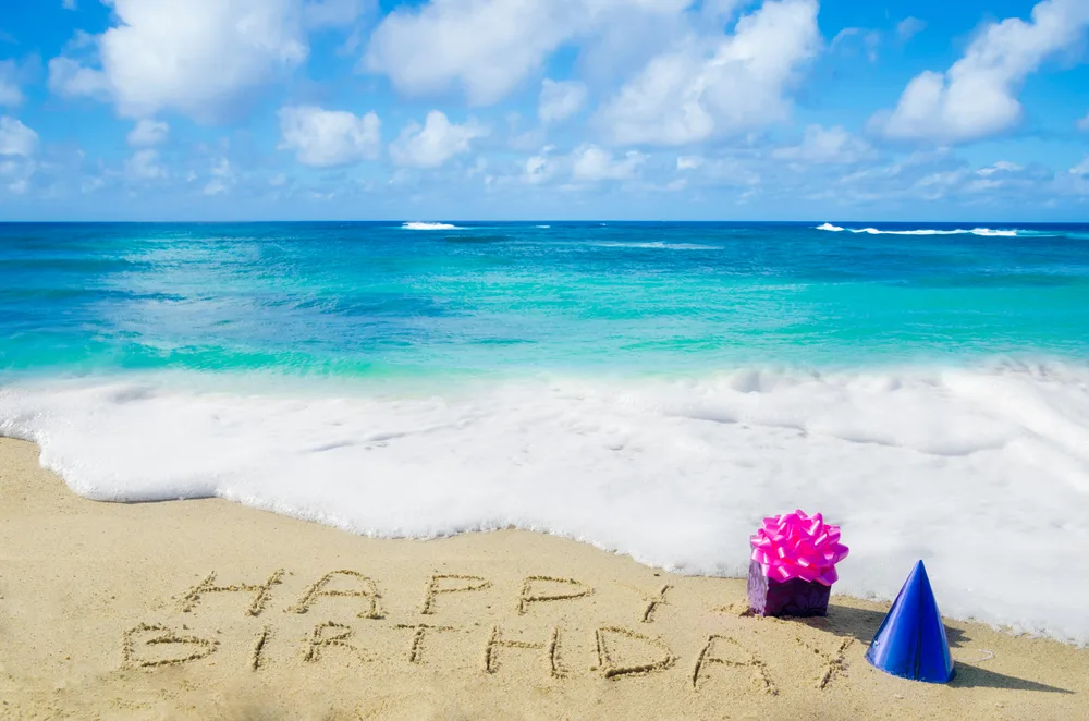 Happy Birthday written in the sand on a tropical beach with party hats for an article showing the 10 best places to go for your birthday trip