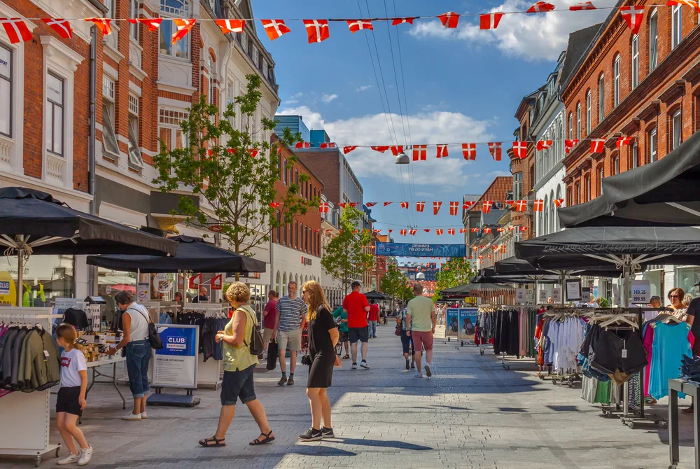 For a guide titled Is Denmark Safe to Visit, a city market in Esbjerg, Syddanmark is pictured on a nice day with flags flying between the tall brick buildings