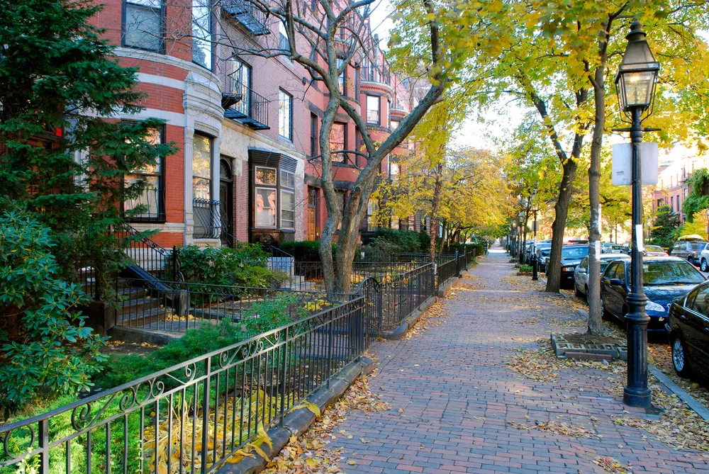 Red brownstone buildings pictured in Autumn, the least busy time to visit Massachusetts, pictured on a street in Boston with yellow and brown leaves falling on the stone path