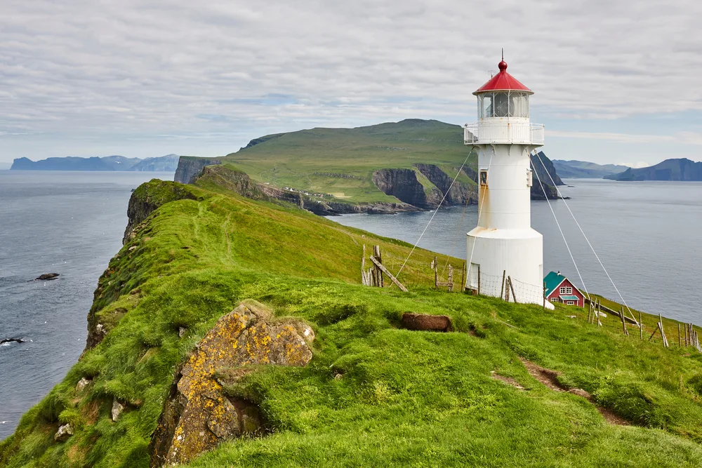 White lighthouse pictured on a rolling hill with steep cliffs on either side on the Faroe Islands, pictured for a guide to whether or not Denmark is safe to visit