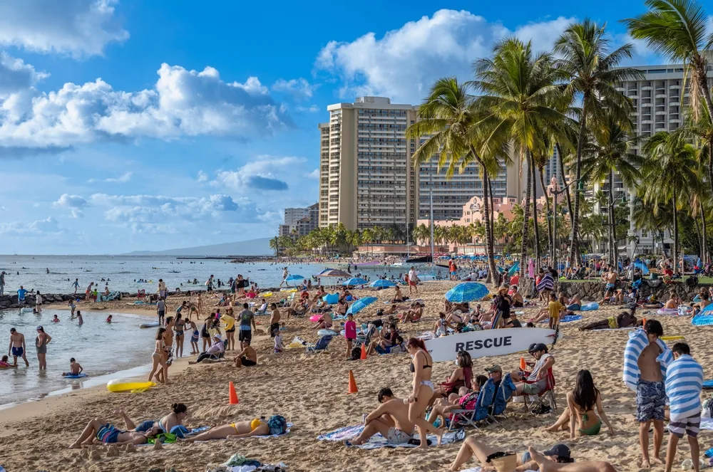 Countless people lying on the beach in the middle of the hotels under a blue sky for a piece on whether or not it's safe to visit Honolulu