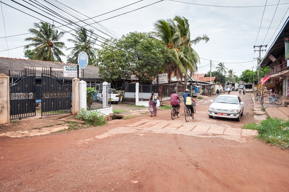 People on the main road in Negombo, Sri Lanka, with semi-wet dirt between old-looking buildings with exposed power lines running under the clouds