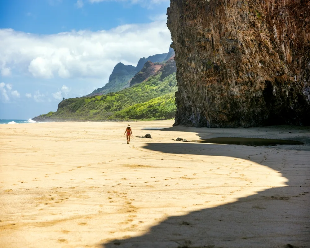 Woman walking along the Na Pali Coast on the Kalalau Trail and being dwarfed by the giant rock cliffs above to show that Kauai is very safe