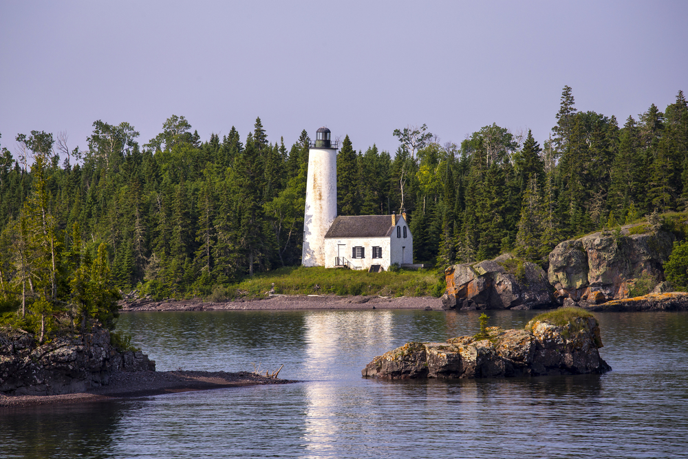 White Rock Harbor Lighthouse pictured overlooking the bay on a hazy day with still water in the front and pine trees in the background