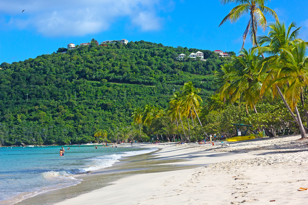 Picturesque tropical beach on St. Thomas pictured with a few people swimming in the water and waves lightly lapping the white sand with palm trees growing over the scene, with a giant hill in the background that's covered in thick vegetation