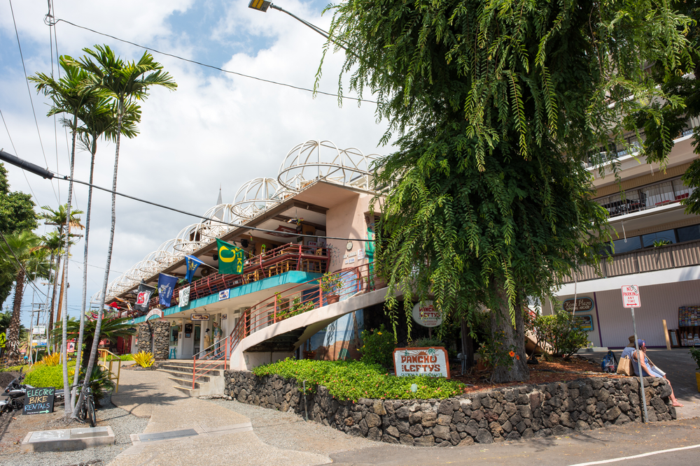 Photo of a market and apartments with colorful flags outside and a black rock wall pictured for a piece on whether the Big Island is safe to travel to