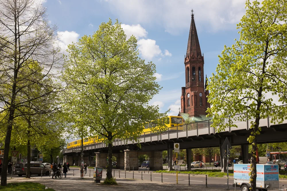 For a guide to whether or not Berlin is safe to visit, a photo of Goerlitzer Park in Belin is pictured on a late-summer afternoon with a yellow tram making its way down the tracks overhead
