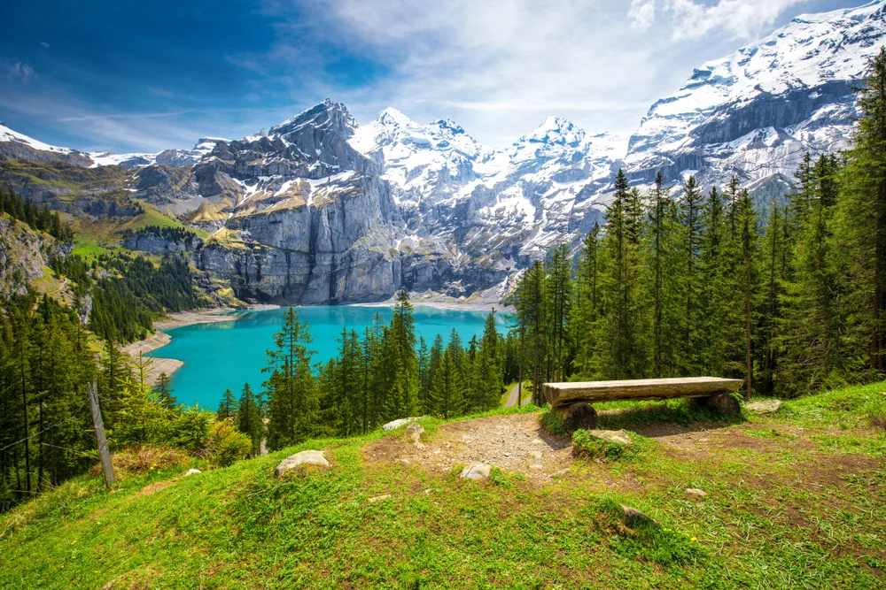 Swiss Alps with a turquoise Oeschinnensee lake and trees with a bench to relax indicates Switzerland's mountains as one of the best bucket list trips