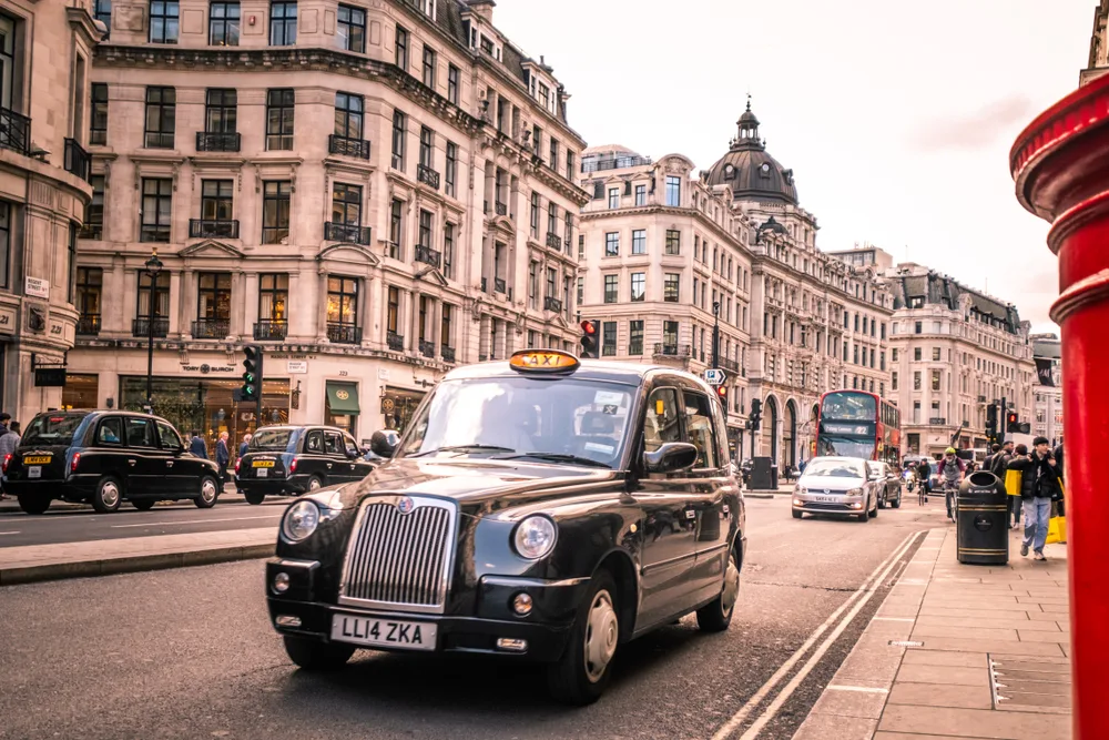 Black London taxi cab on the West End for a guide to the average trip to London cost with hotels on either side and traffic on the streets