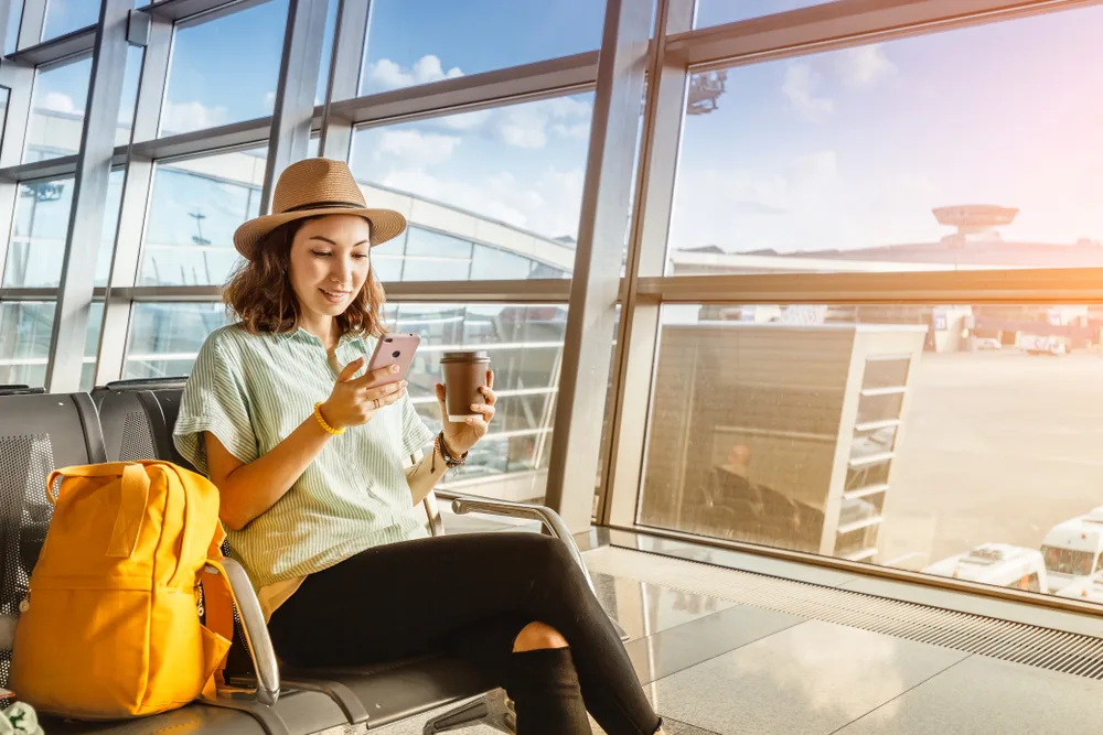 Young woman sitting in an airport with her yellow luggage next to her uses her smartphone to confirm her flight details on Google Flights