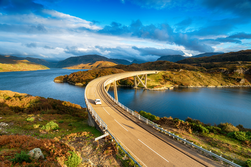 For an image for a guide to the average Scotland trip cost, a photo of a white car driving on the left side of the road over the Kylesku Bridge