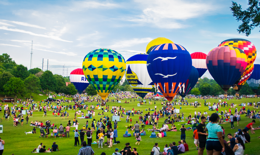 A bunch of colorful hot air balloons pictured floating above the green park grass during the summer, the hottest time to visit Atlanta for a piece on whether or not it's safe to visit