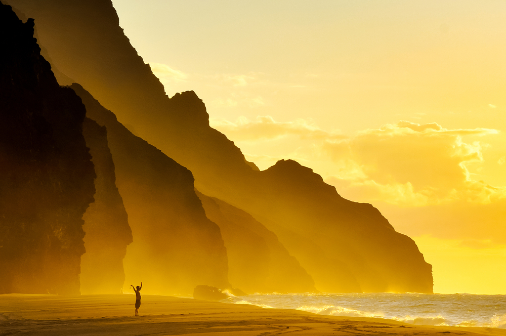 Woman throwing her hands in the air in an artistic-style image on Kalalau Beach in Kauai with an orange hue, as seen at sunset with cliffs in the background for a guide to the average trip to Hawaii cost