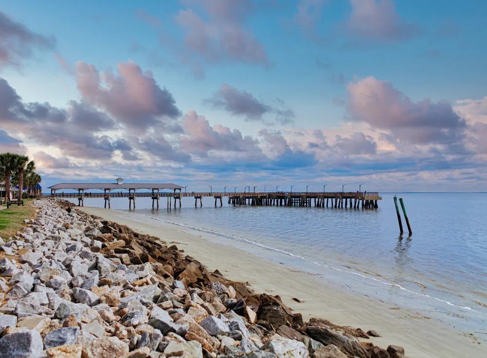 Old wooden fishing pier in St. Simon's Island, GA with clouds in the sky and a rock seawall behind the sand for a list of the best beach vacation ideas in the US