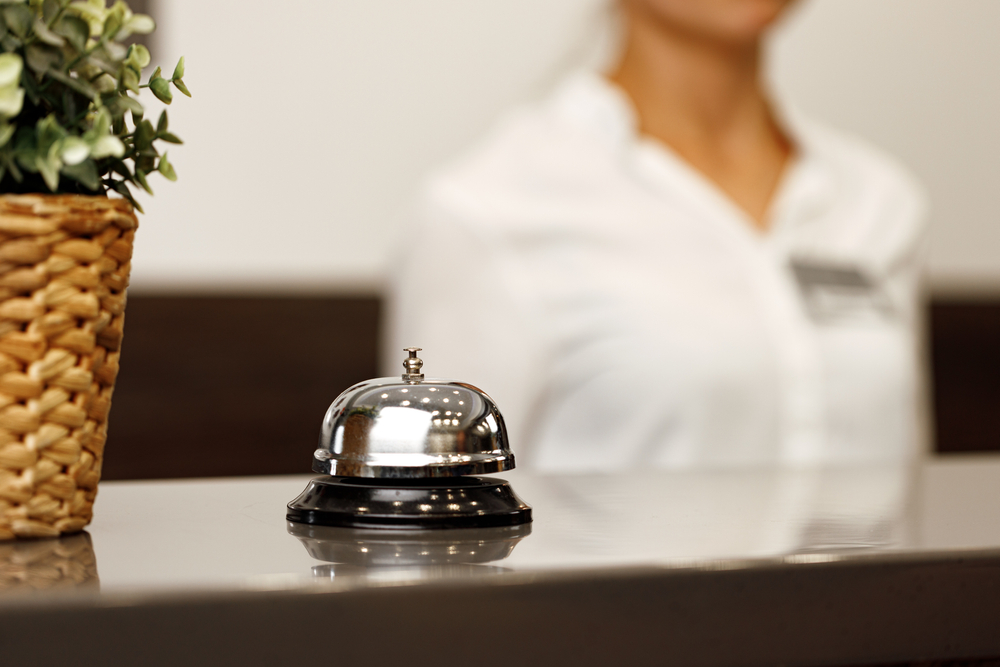 Hotel service bell in focus with front desk worker in a white button-down shirt blurry behind it for a piece asking can you pay for a hotel in cash