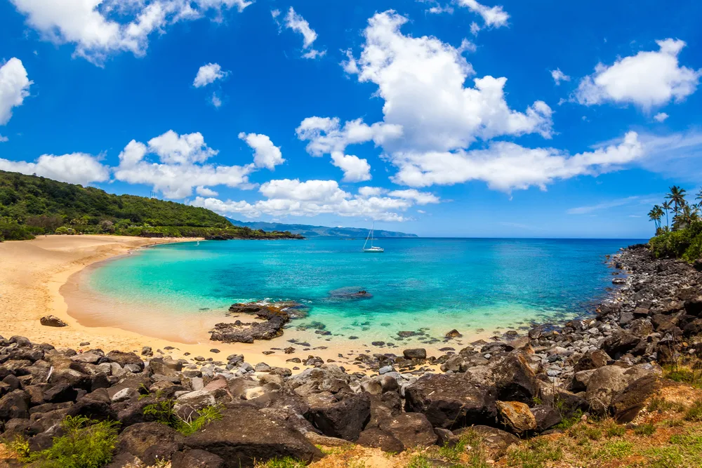 View of Waimea Bay Beach in Maui, Hawaii with fluffy clouds overhead and rocks along the shore for a list of the best beach vacations in the US