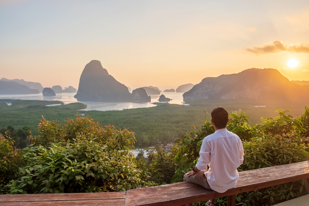 Guy relaxing alone on the edge of a railing overlooking Phang Nga Bay as an image for a guide titled What Trips to Thailand Cost on Average