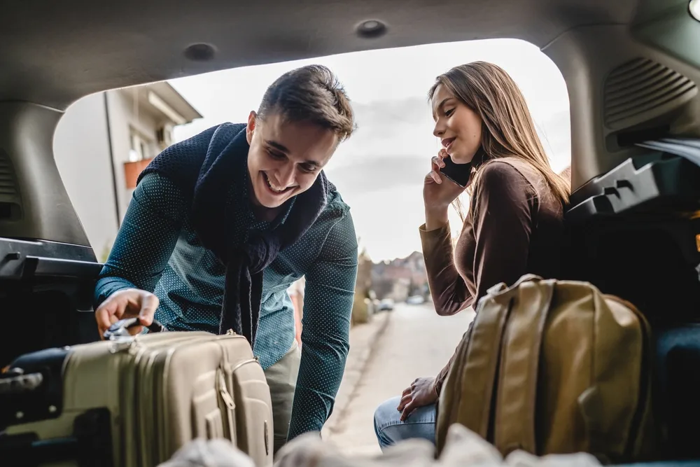 Young couple smiling and loading luggage into the back of a rental car as they're about to go out of state