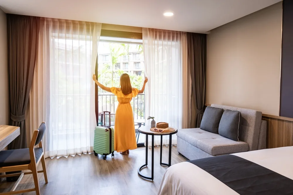 Female traveler in an orange dress opens the balcony curtains in her room with luggage beside her for a piece answering the question, can you pay for a hotel in cash?