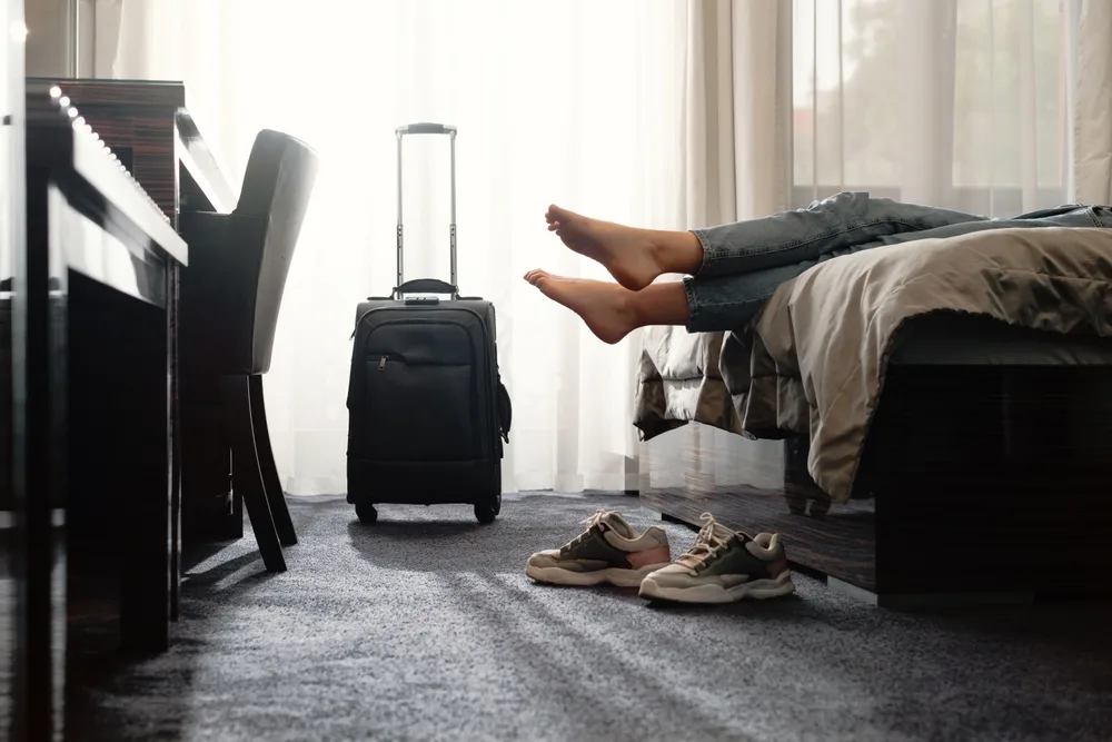 Woman who learned how to get free hotel rooms rests with her shoes off on the hotel bed after bringing her luggage to her room as her vacation begins
