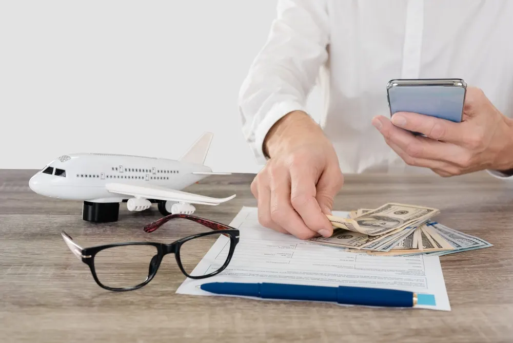 Man holding smartphone touches money on his desk with a model plane, glasses, and documents as he wonders why are flights so expensive