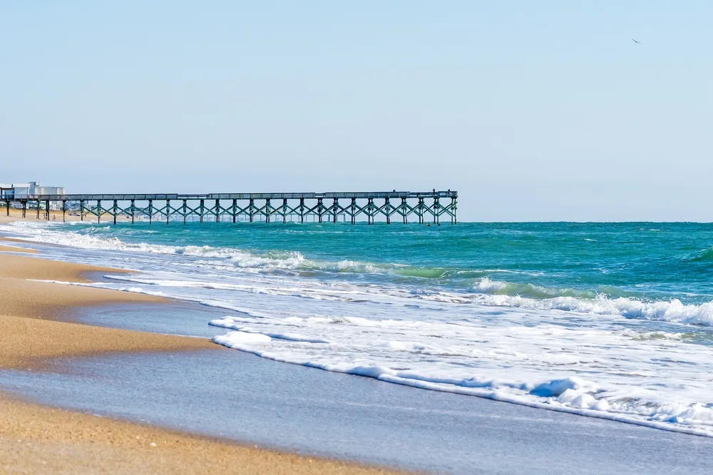 Wrightsville Beach in Wilmington, NC statistical area shown with a pier and gentle waves on a sunny day as one of the best East Coast vacations spots