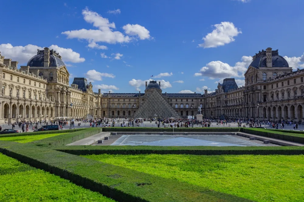 Gorgeous sunny day with the Louvre and its glass and metal pyramid out front with green grass all around pictured for a guide titled What Does a Trip to Paris Cost
