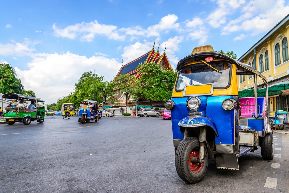 For a guide titled what does a trip to Thailand cost, a blue tuktuk pictured sitting outside of the historic temple in Chiang Mai
