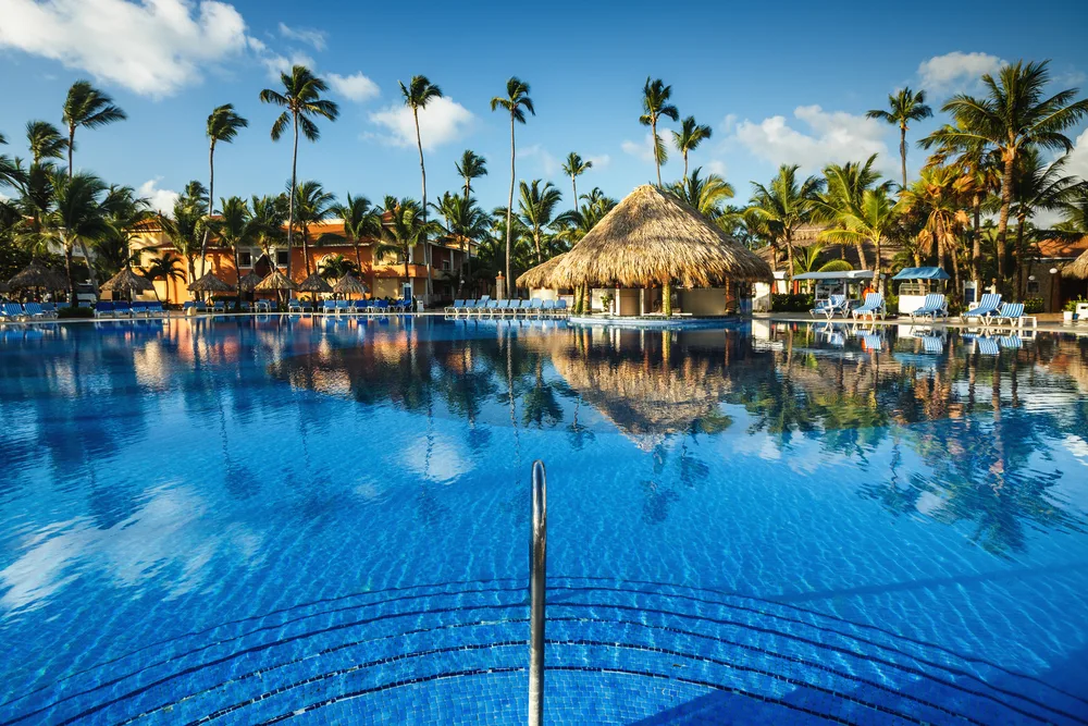 Empty tropical swimming pool at a luxury resort overlooking thatched huts and palm trees on a beautiful day for a Puerto Rico vs the Dominican Republic cost comparison