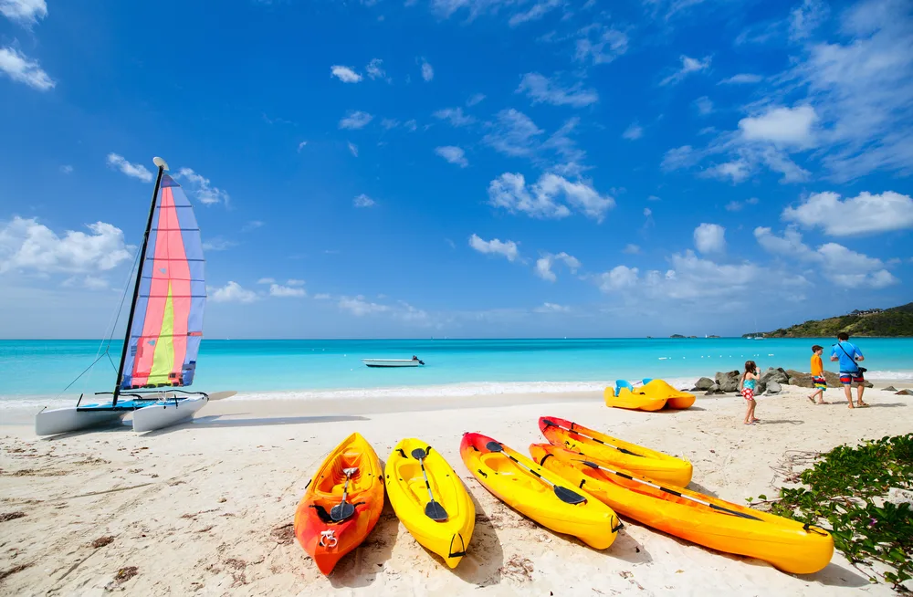 View from an Antigua beach with kayaks and wind surfing board on the sand as kids play off to the side for a list of the best family vacations for all ages