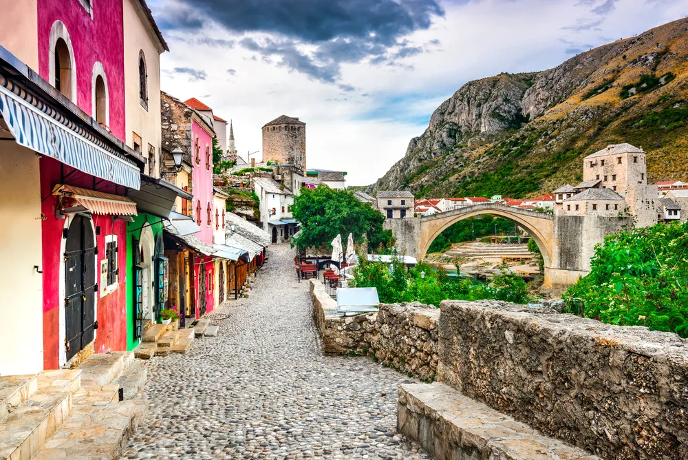 Picturesque view of a cobblestone street in Mostar, one of Bosnia's best areas to stay, with its famous bridge over the river and colorful home fronts facing the river