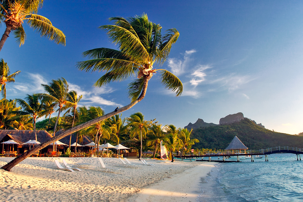 Gorgeous scene with over-water bungalows and a dock above teal water with a white-sand beach to the left for a guide to what a Bora Bora trip costs