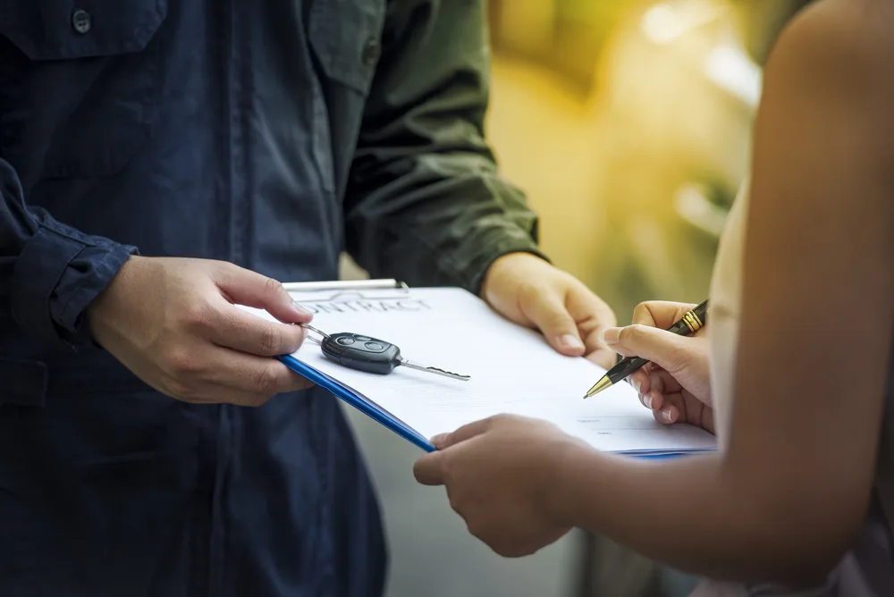 Woman signing a rental car agreement on a clipboard as the salesman holds the car key for a piece asking can you take rental cars out of state?