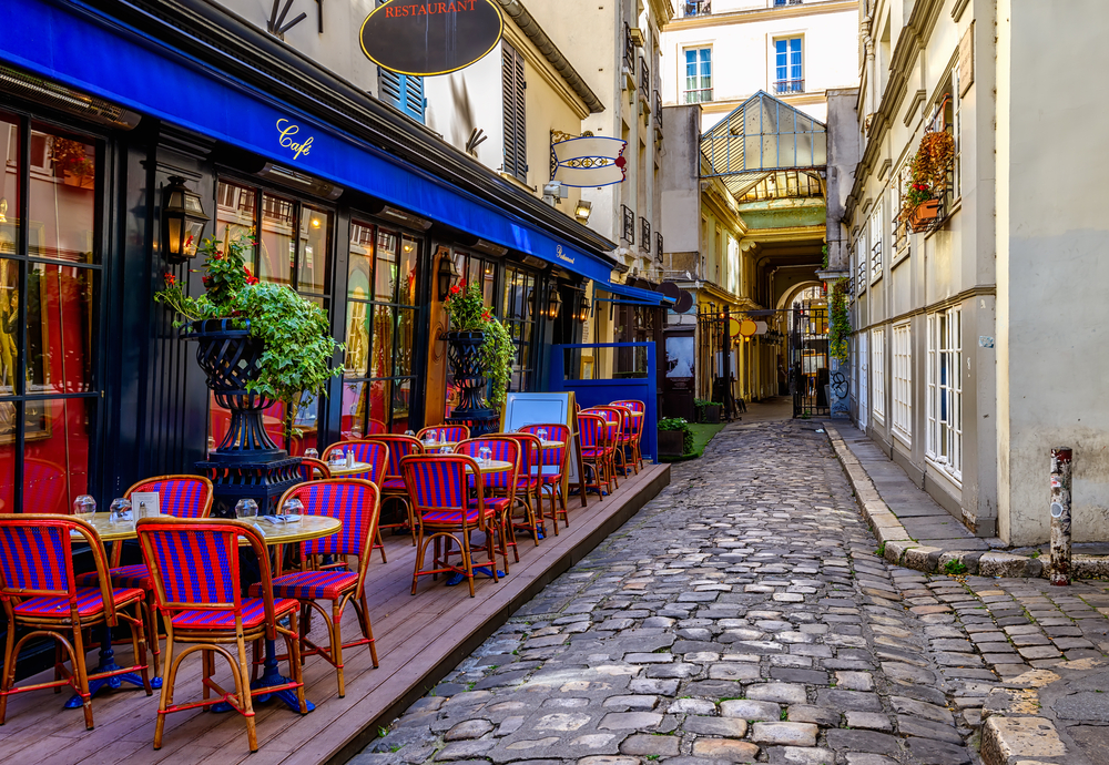 Neat little cafe in Paris with red chairs outside on the patio and a cobblestone street alongside it for a guide to the average trip to France cost