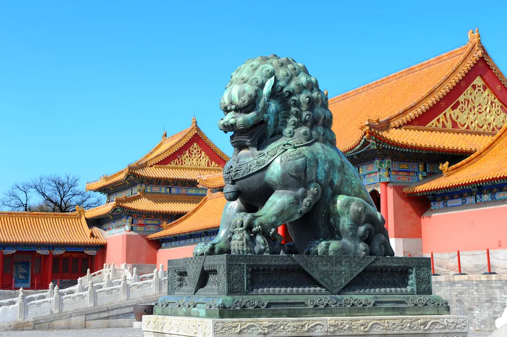 The Forbidden City in Beijing is guarded by a large lion statue with ancient royal palaces inside the old walled city, making it one of the great bucket list travel ideas