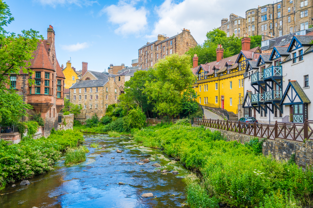 Sunny day over Dean Village in Scotland for a guide to the average trip cost, depicting an old Bavarian-styled town on the water's edge