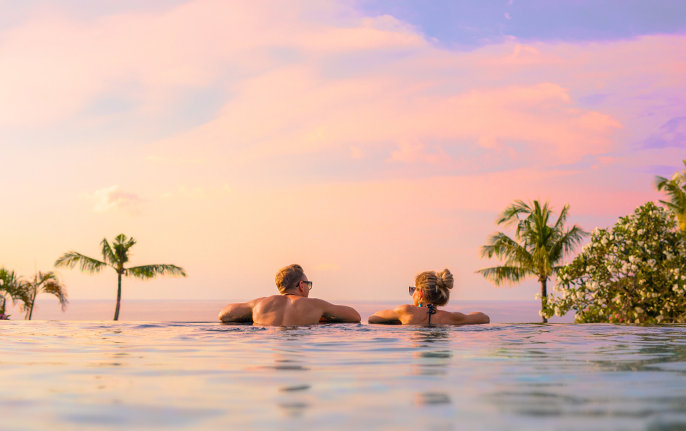 Couple looks lovingly at each other at sunset in an infinity pool overlooking the ocean in a tropical getaway for a honeymoon with palm trees in the distance