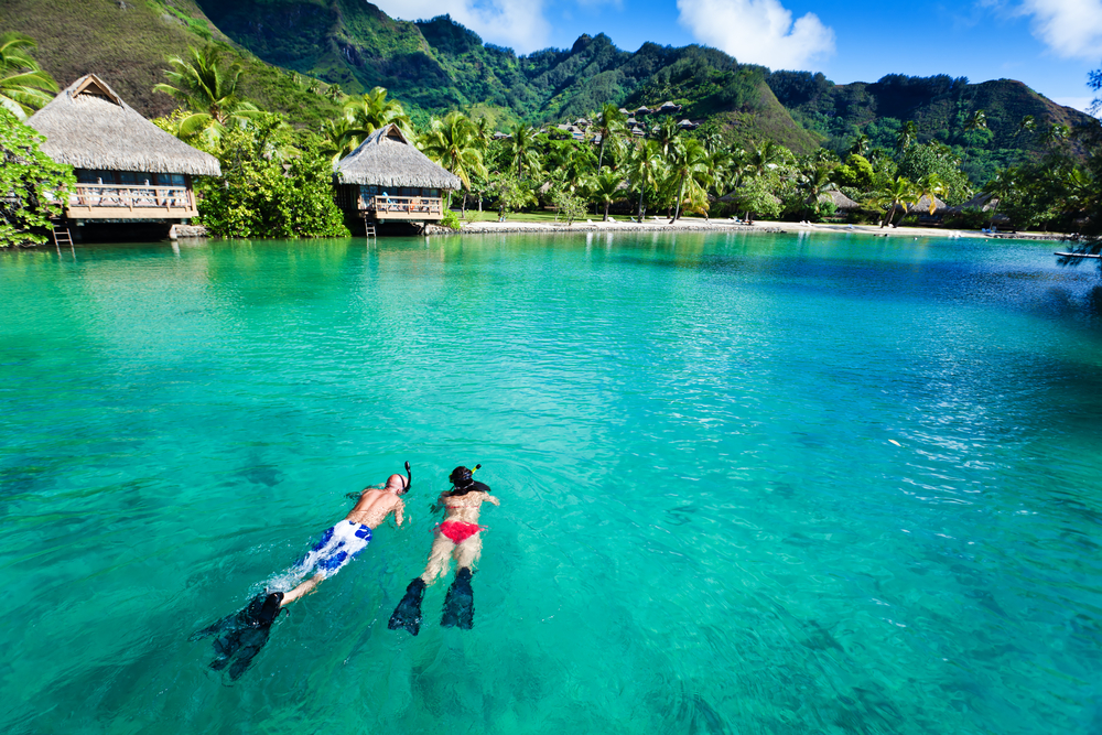 Newlywed couple snorkeling in clear ocean water next to overwater bungalows to indicate where you should go for a honeymoon
