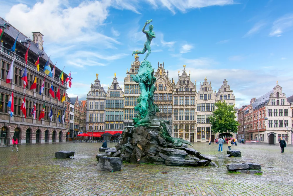 Statue of a naked man holding a hand in throwing position, while standing above an object being held by other statues, in the middle on an open area surrounded by old structures, an image for a travel guide about trip cost to Belgium.