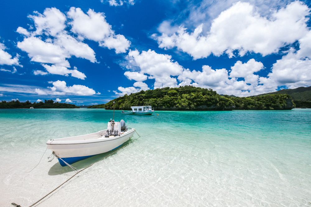 A boat docked on the shallow part of a beach with clear water and white sand, and small island filled with trees can be seen in background. 