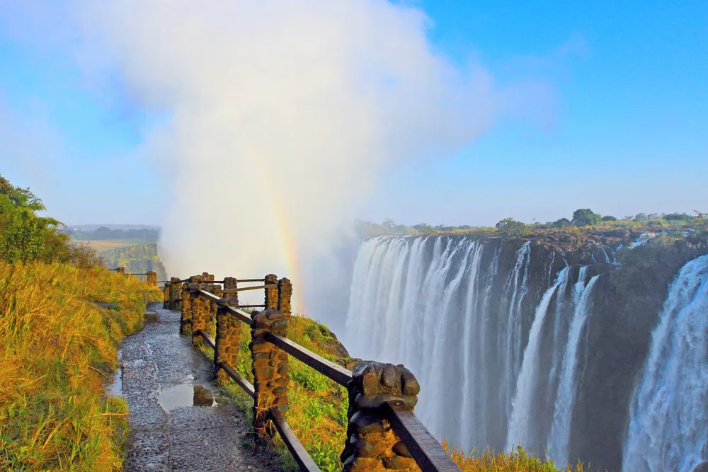 A close view of a large falls on the opposite side with wooden railings and a rainbow can be seen from the moisture from the falls.