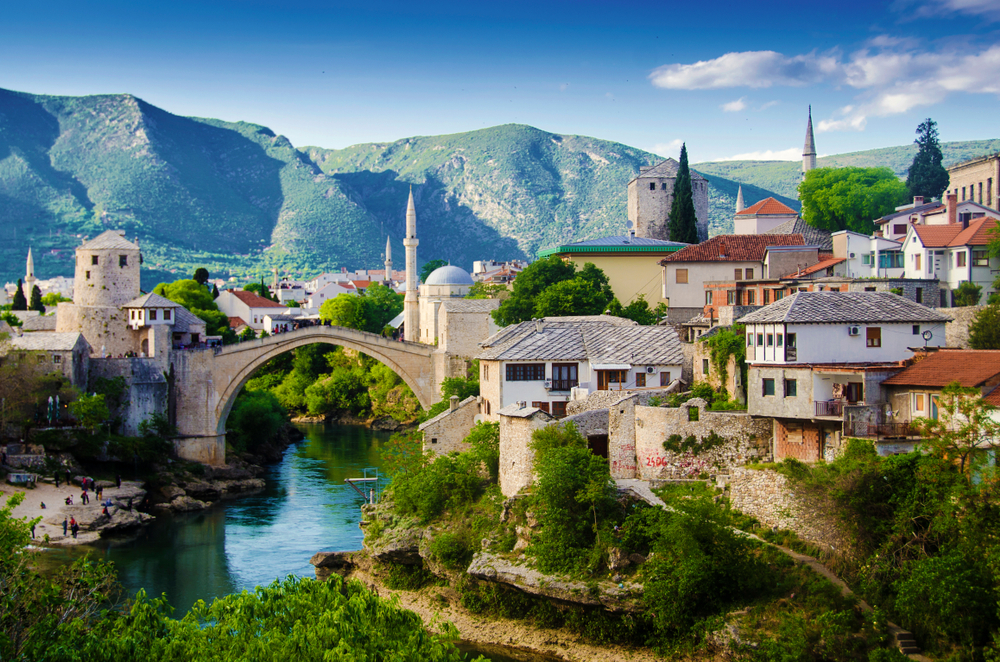 Mostar in Bosnia and Herzegovina shown with mountains in the background and an old bridge leading over a river through the town on a list of the cheapest places to visit in Europe