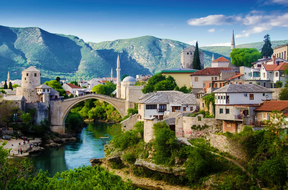 Mostar in Bosnia and Herzegovina shown with mountains in the background and an old bridge leading over a river through the town on a list of the cheapest places to visit in Europe