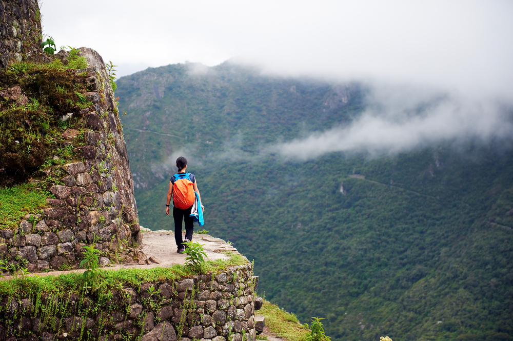 A woman walking on a trail by the side of a mountain, captured for a piece on an article about trip cost to Peru, in background is a foggy mountain covered in lush trees.