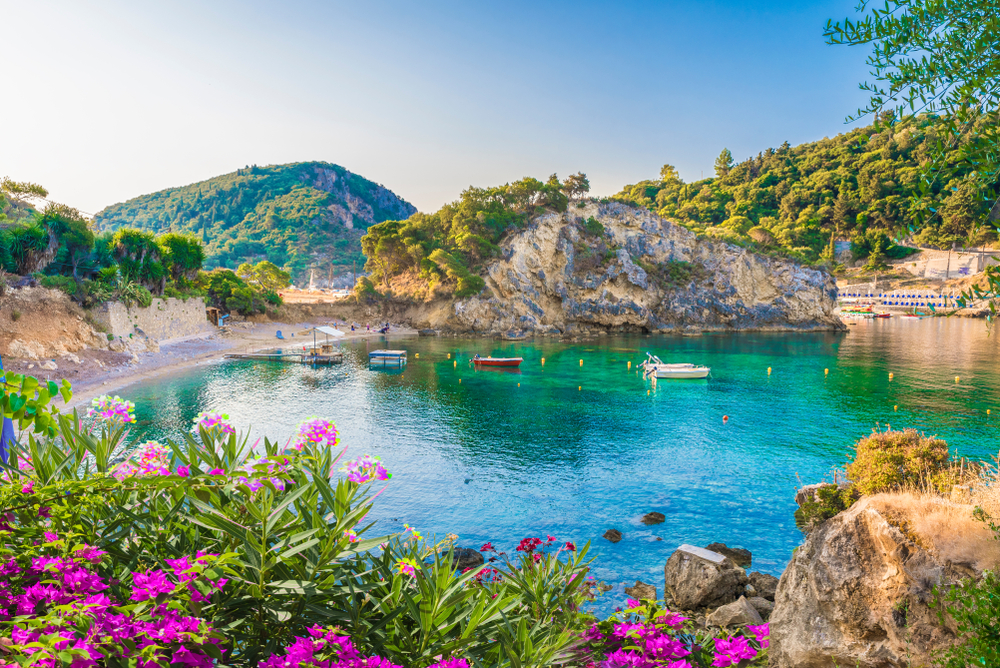 Paleokastritsa Bay in Corfu, Greece with flowers in bloom in the foreground and beautiful water with green hills surrounding it for a list ranking the top party islands in Greece