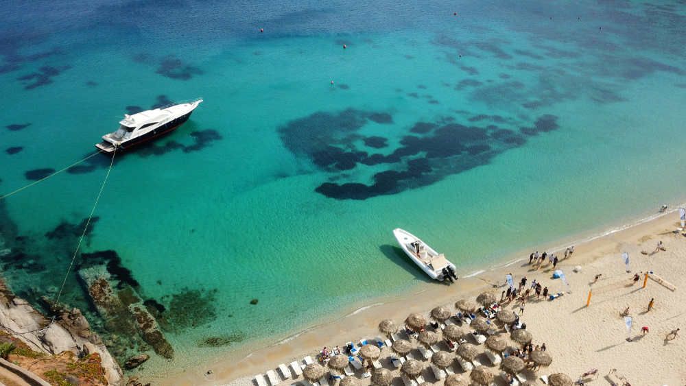 Aerial view of a beach with tourist sunbathing and others enjoying the shore where a boat can be seen moored a little farther to the shore, an image for a travel guide about safety in visiting Mykonos.