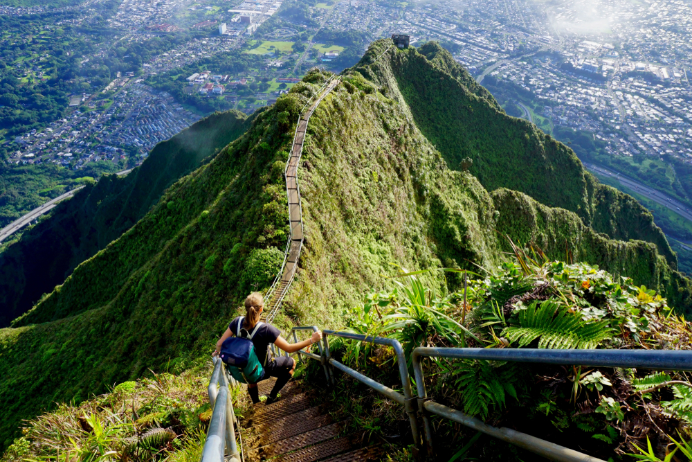 Haiku Stairs at the Stairway to Heaven in Oahu, Hawaii shown from above as a hiker makes their way down with a backpack for a guide to traveling between the Hawaiian Islands