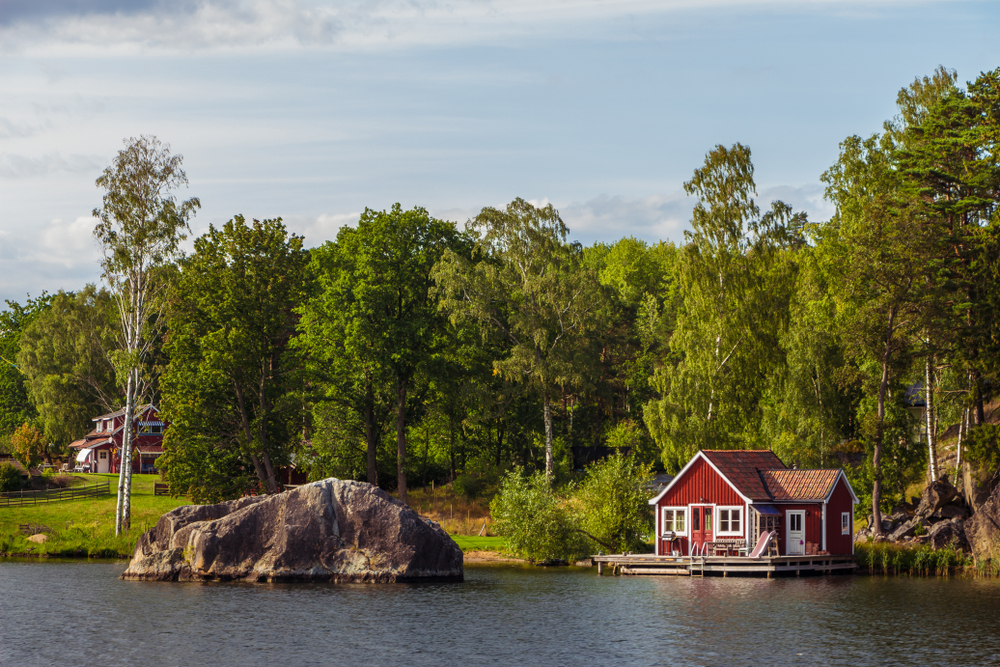 A small cabin by the lake with a large rock beside it, and another small cabin can be seen at a distance surrounded by trees.