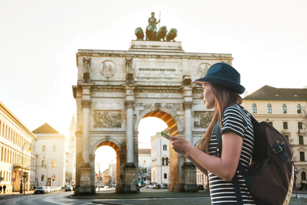 A woman wearing stripes and a hat holding her phone while scanning the surroundings of the city, and in back ground is the historic Triumphal Arch, an image for a travel guide about safety in visiting Munich.