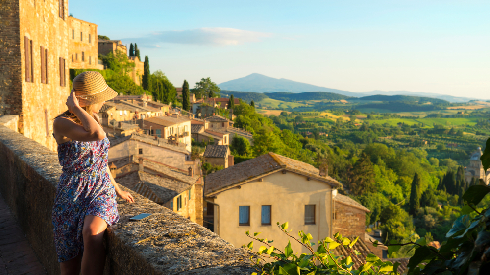 A woman sitting on the edge while looking at the Tuscan landscape while holding her hat during a sunset. 
